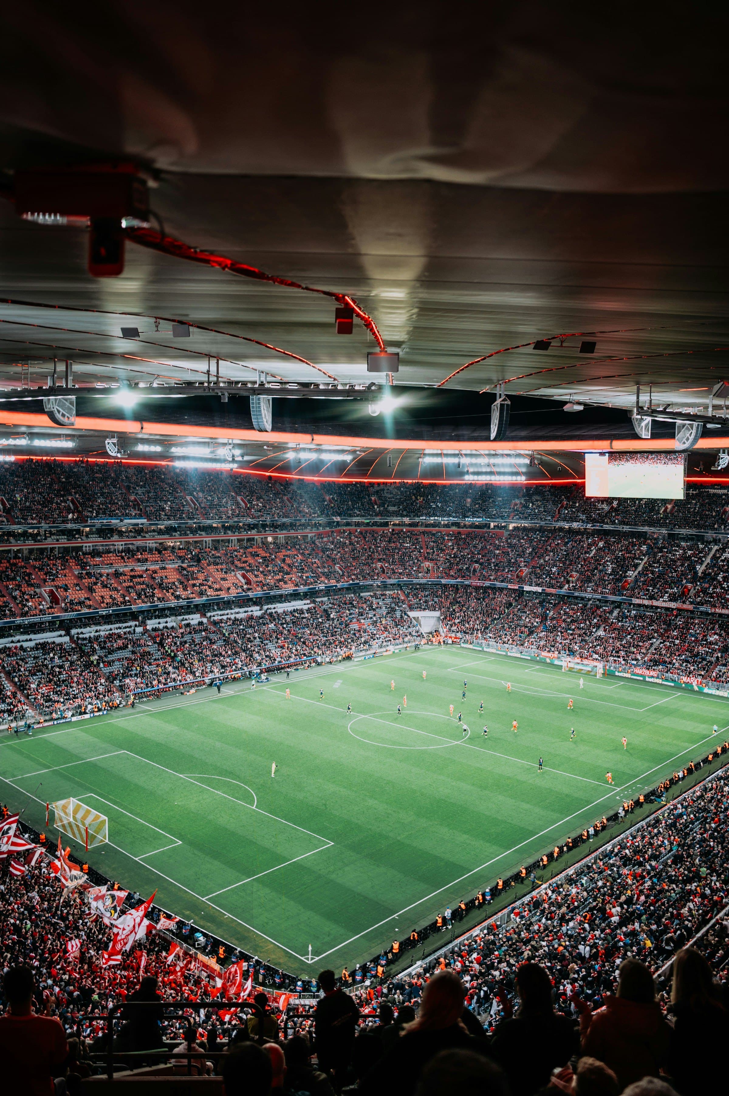 A floodlit football stadium at night, packed to the roof, flags in the stand